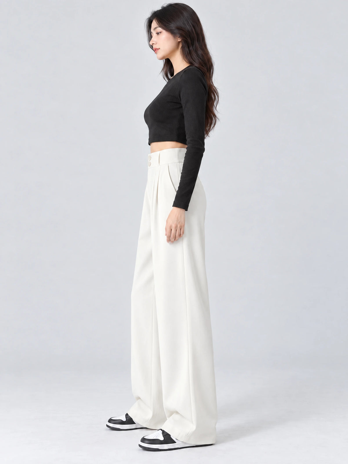 Side studio view of a model wearing White high waist wide leg pants with a buttoned waistband and pleated front.