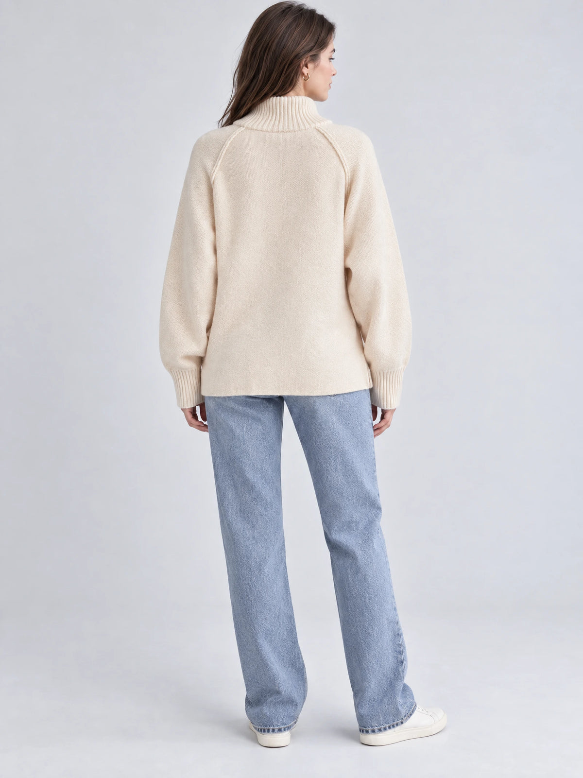 Back view of a model wearing a Beige knit full zip sweater with a ribbed high collar, raglan sleeves, and ribbed cuffs.