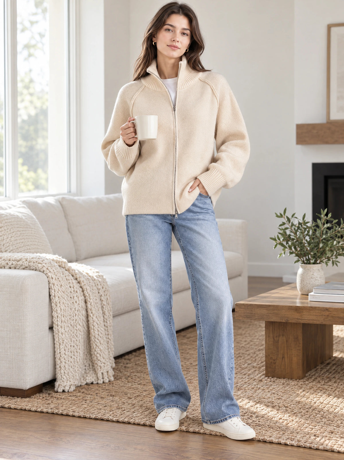 Model wearing a Beige knit full zip sweater with a ribbed stand collar while holding a mug in a sunlit living room.