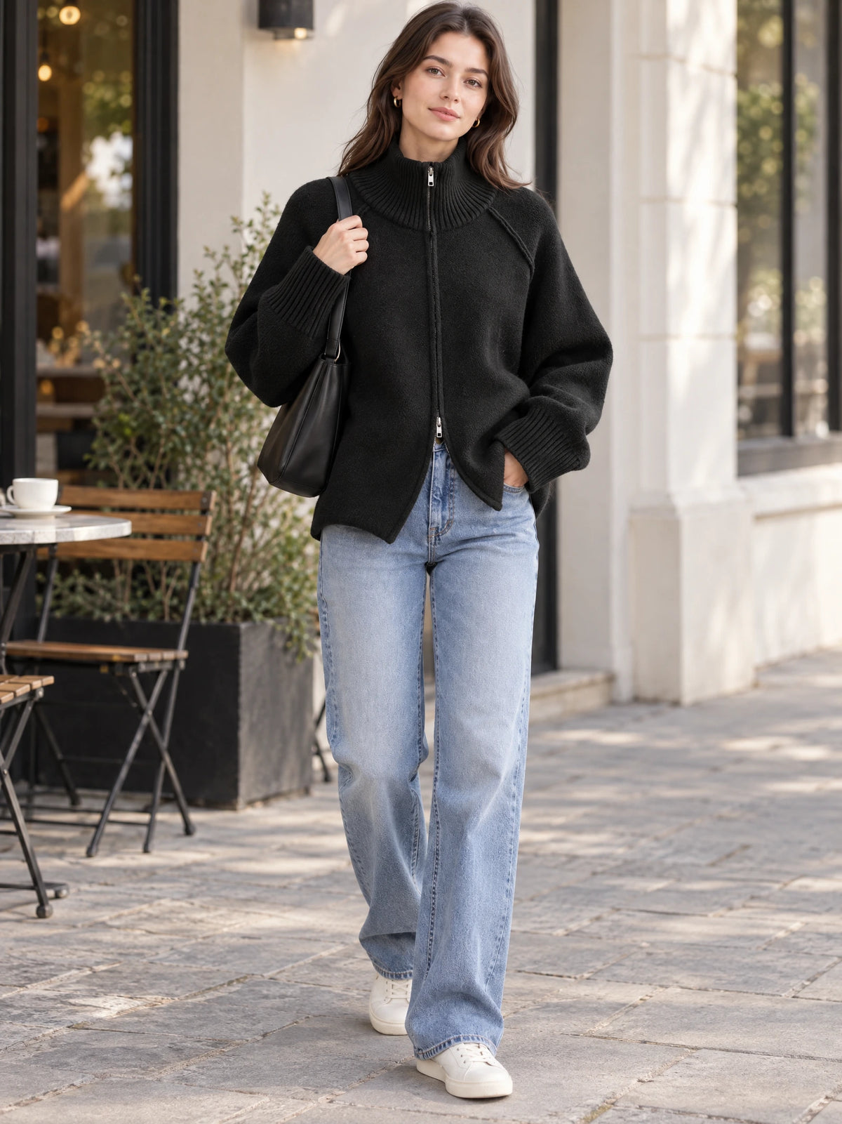 Model wearing a Black knit full zip sweater with a ribbed high collar and cuffs while walking past a sidewalk café.