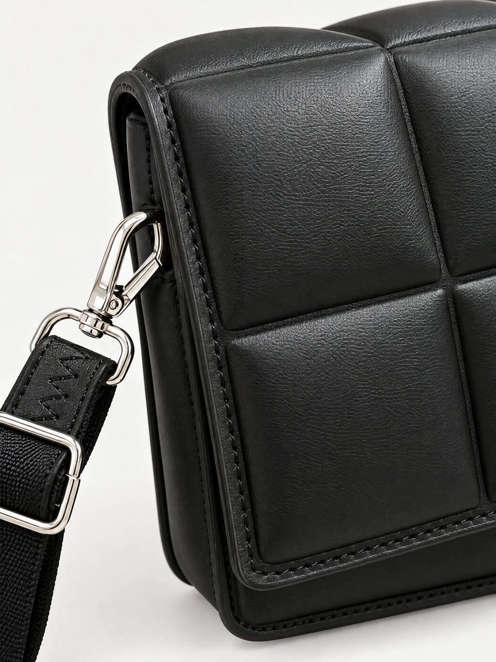Close-up of a Black leather cross body bag showing textured panel stitching and a strap clasp.