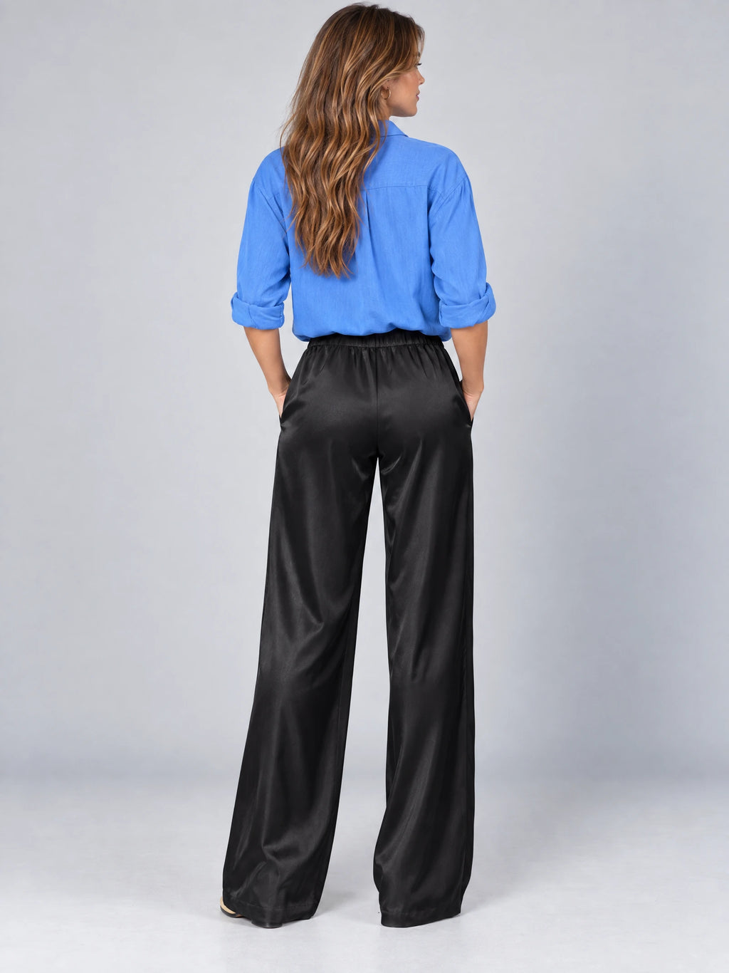 Rear studio view of a model wearing Black satin wide leg pants with an elasticized high waist and flowing full-length legs.