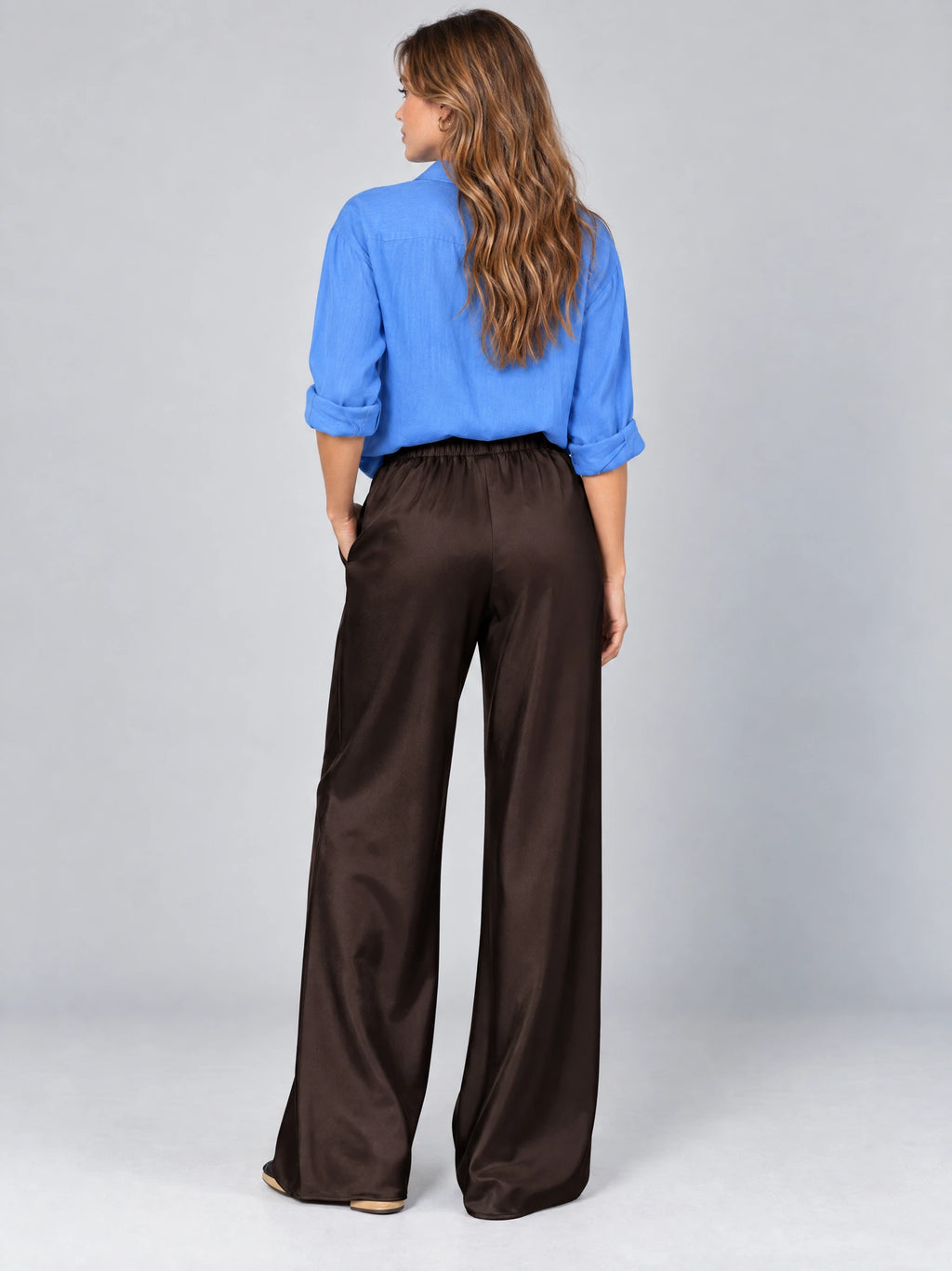 Rear studio view of a model wearing Brown satin wide leg pants with an elastic high-rise waistband.