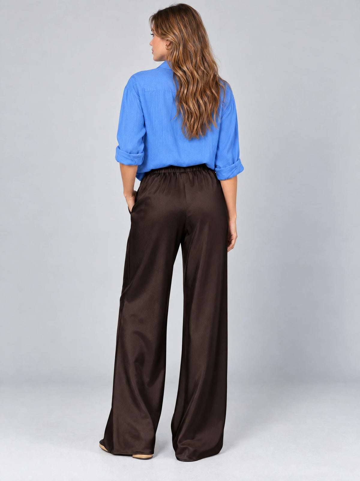 Rear studio view of a model wearing Brown satin wide leg pants with an elastic high-rise waistband.