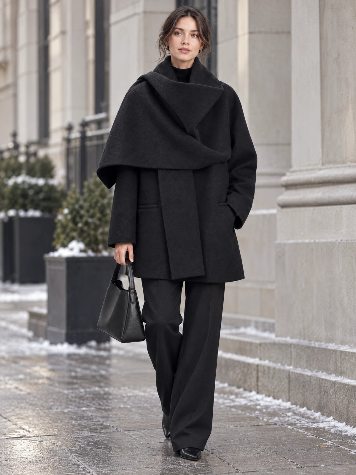 Model walks along a snowy city sidewalk in a Black scarf-collar wool coat, with one hand in a pocket and a handbag at her side.