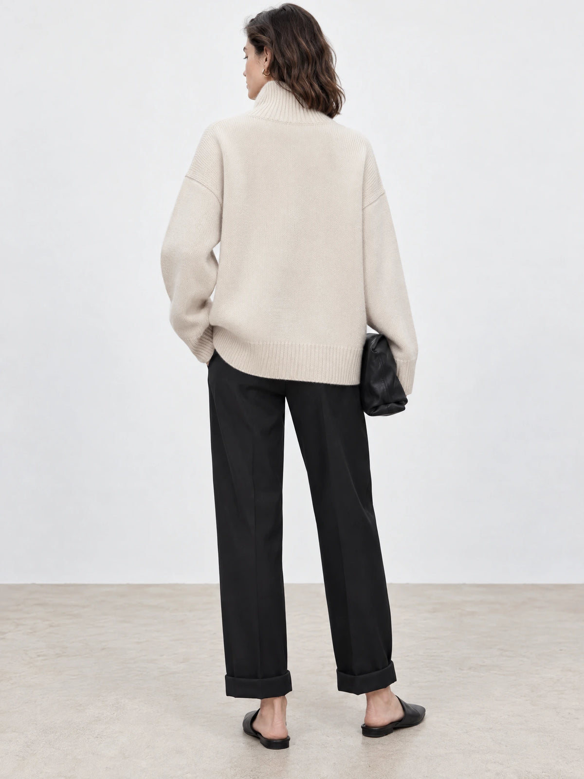 Rear view of a model wearing a Beige wool turtle neck sweater with a ribbed high collar, relaxed sleeves, and ribbed hem.